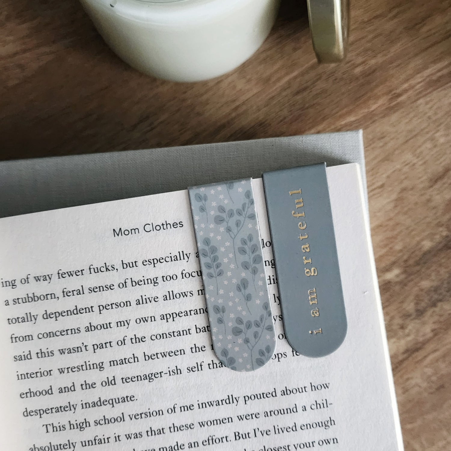 An open book with two bookmarks, one floral and one gray with gold text reading i am grateful, on a wooden table beside a candle.