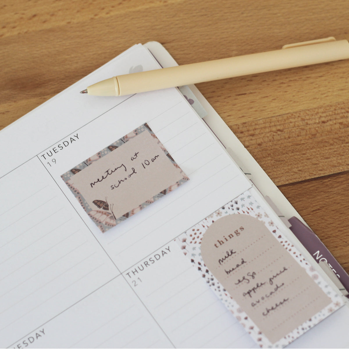 A Florals Mini Sticky Note from Maylay Co., highlighted with delicate floral patterns, displays a handwritten message saying "meeting at 10 am on Tuesday." Beside it, a pen lies next to another sticky note labeled "things," which includes items like milk and bread. The wooden surface beneath them offers a warm contrast in the background.